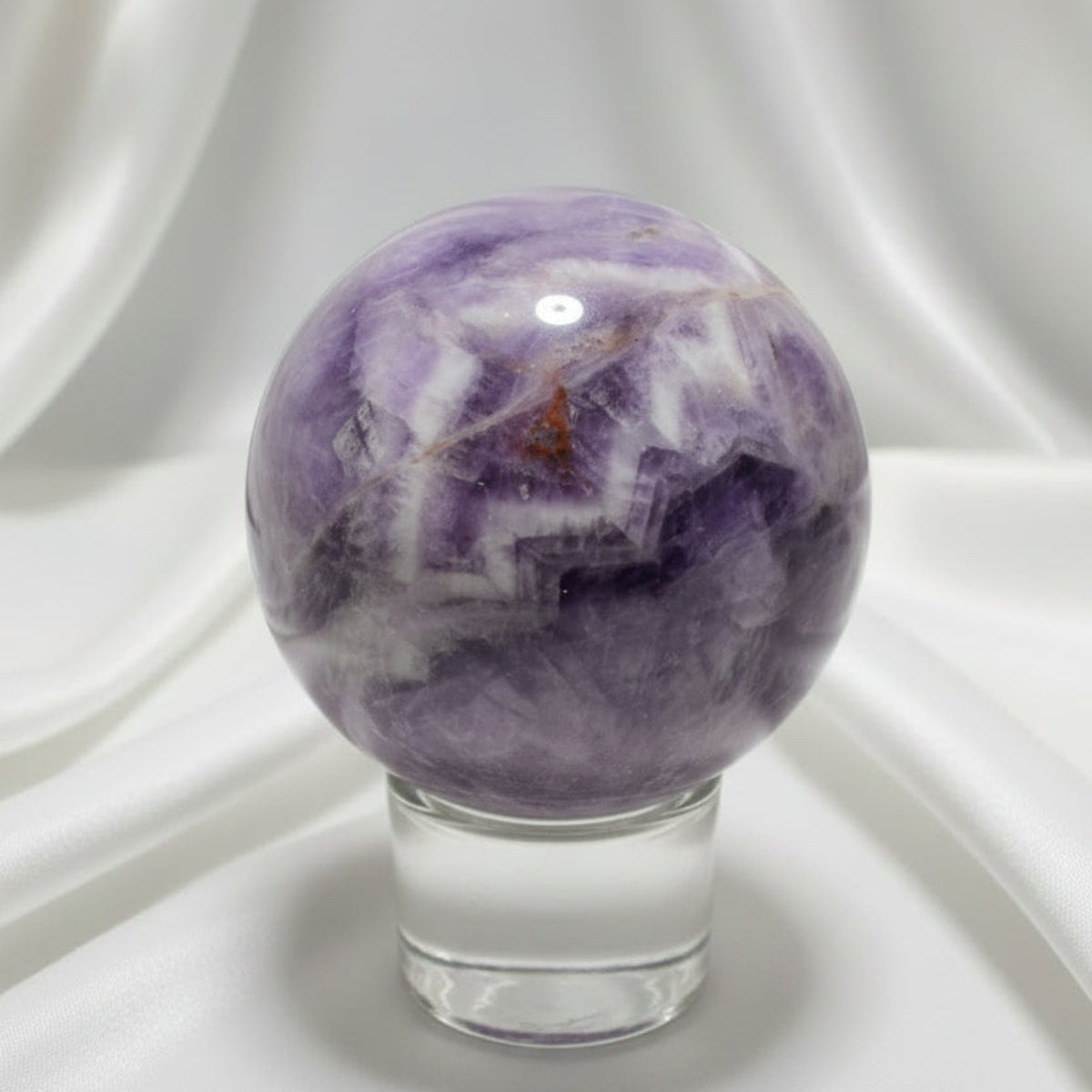 Purple, White, clear chevron amethyst carved sphere with iron inclusions on a glass stand with a white fabric in the background