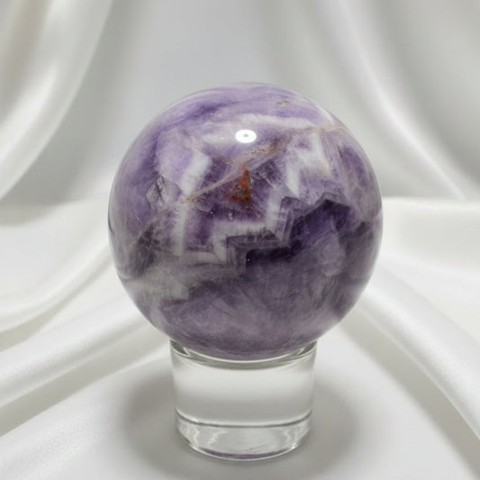 Purple, White, clear chevron amethyst carved sphere with iron inclusions on a glass stand with a white fabric in the background