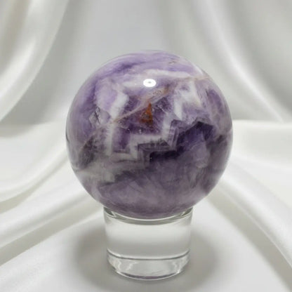 Purple, White, clear chevron amethyst carved sphere with iron inclusions on a glass stand with a white fabric in the background