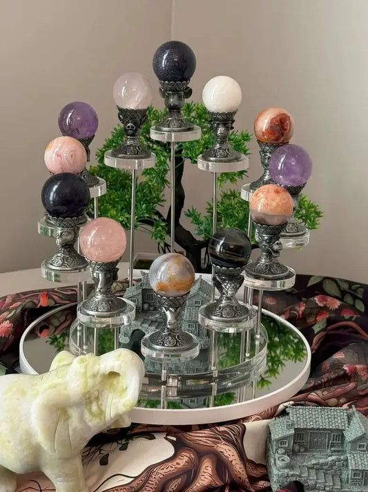 A crystal sphere display stand with multiple polished gemstone balls in different colors, arranged on a mirrored circular base with a green decorative tree behind it.