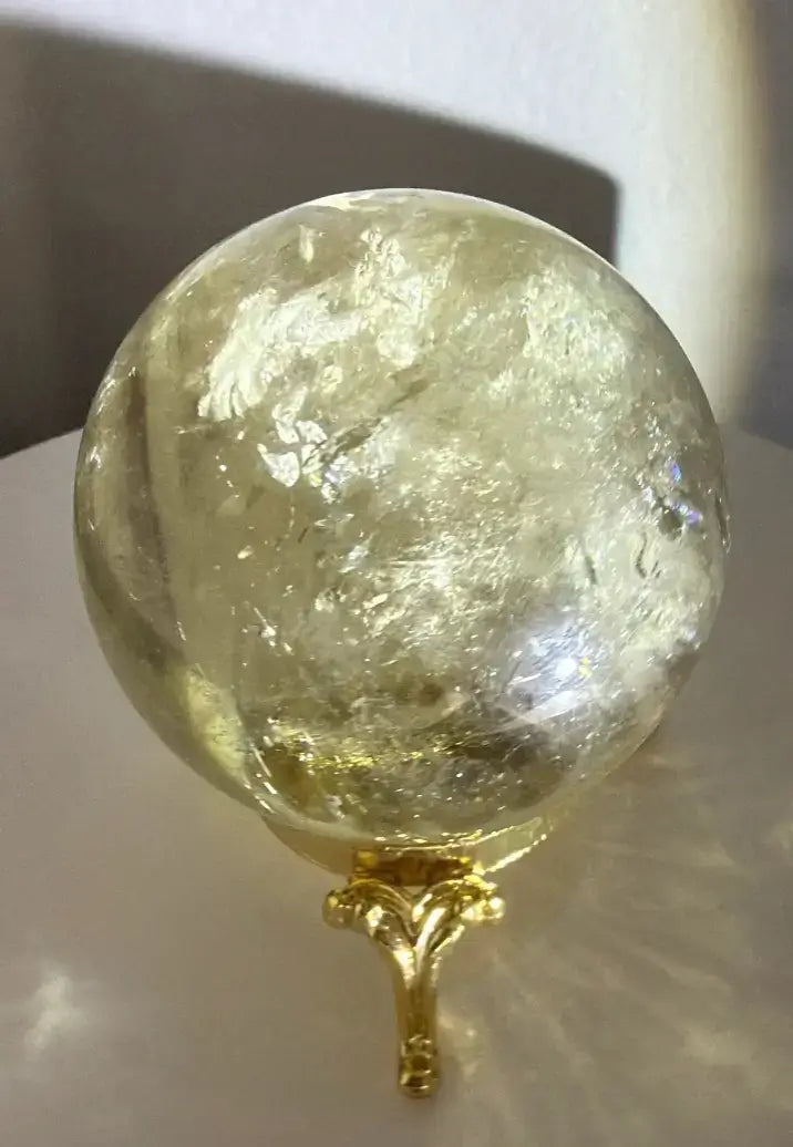close up of a citrine sphere on a gold stand with a shadow background. photo shows the natural yellow hues of citrine and the natural fractures and inclusions
