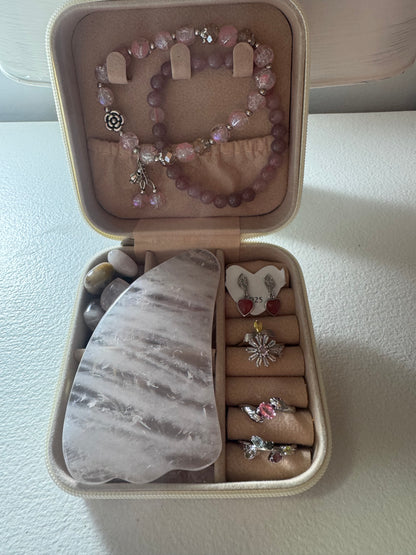 An open jewelry box displaying rings, earrings, bracelets, tumbled stones, and a gua sha.
