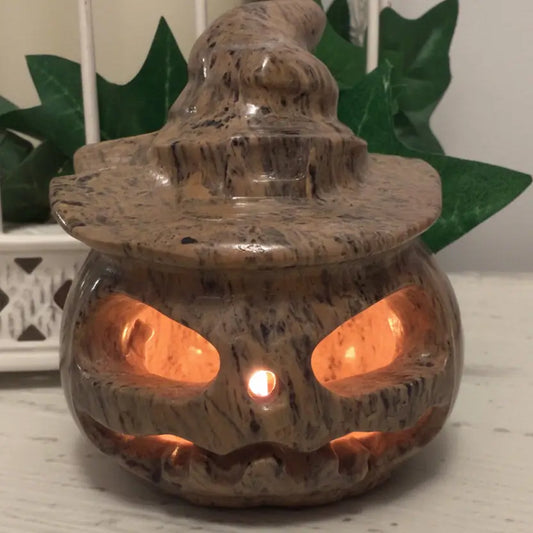 Dendritic Jasper Jack O’Lantern Carving sitting on a white table with greenery behind and candle light inside