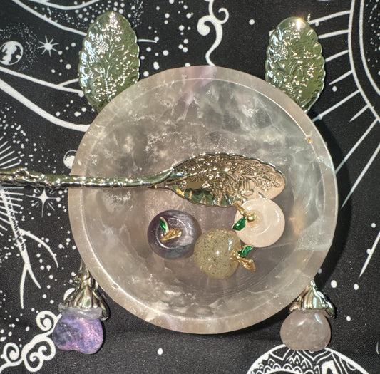 Overhead shot of a crystal bowl containing apple-shaped stones, silver spoons with floral designs on a black and white patterned cloth.
