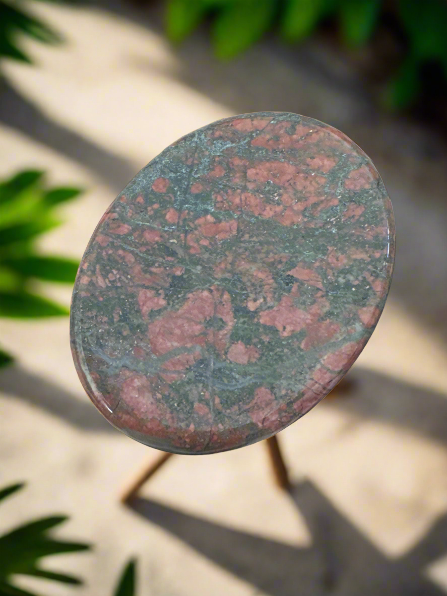 Close up of a small, oval marble table with a red and green speckled pattern, set on a three-legged wooden stand, against a blurred background of plants and sunlight.