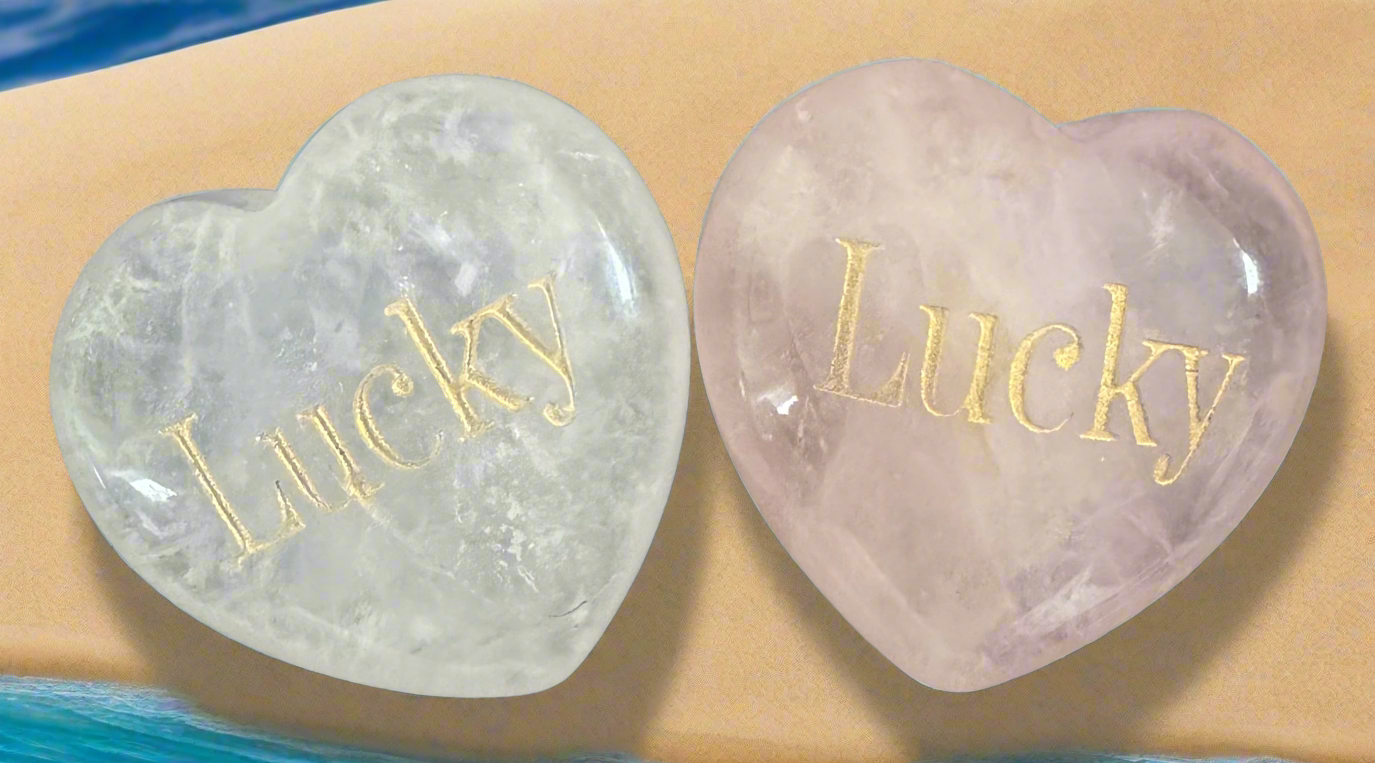 Two heart-shaped crystals, one translucent white and one pale pink, both inscribed with the word "Lucky" in gold lettering, on a beige surface with blue accents.