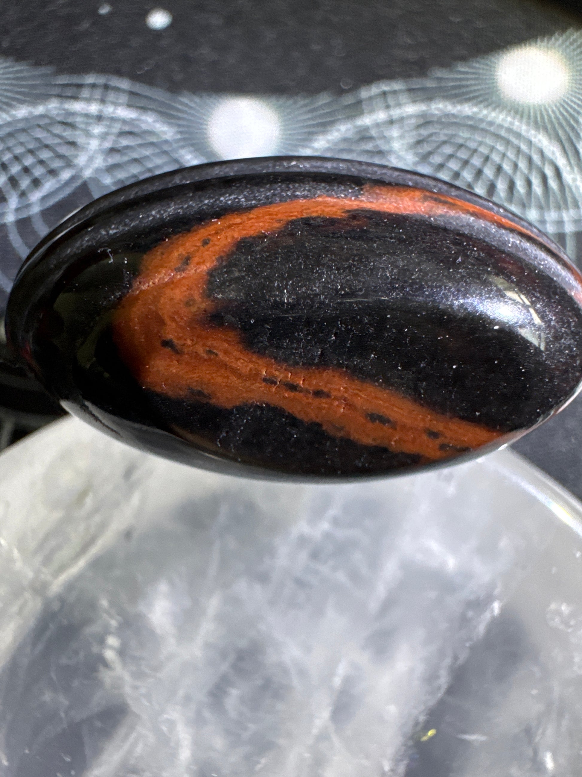 Oval polished stone, black with orange band, resting on a clear surface.
