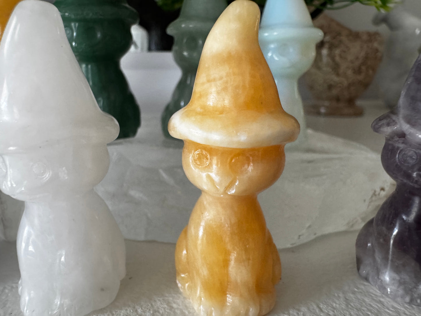 Close-up of a collection of crystal gnomes in different colors: white, orange, green, blue, and purple. The gnomes have hats and small facial features. They are on a white surface with other objects in the background.
