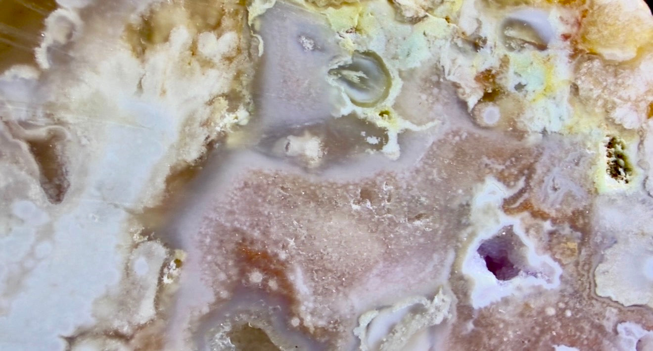 A close-up of a polished flower agate   swirling bands of cream, beige, tan, light brown, and small areas of pale yellow and a faint purple inlay. Hint of a face top right