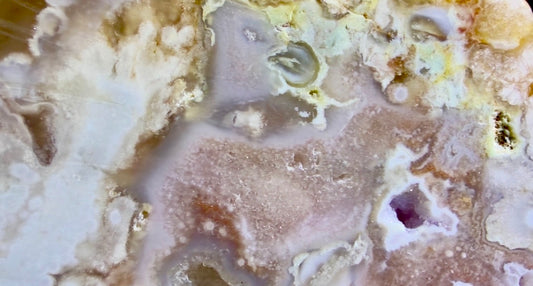 A close-up of a polished flower agate   swirling bands of cream, beige, tan, light brown, and small areas of pale yellow and a faint purple inlay. Hint of a face top right