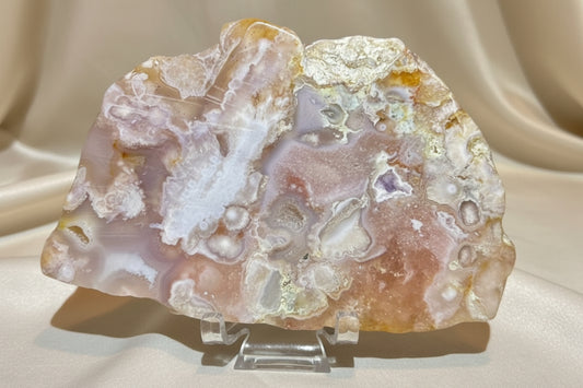 A polished flower agate stone specimen with swirling bands of white, peach, pink, and iridescent gold, displayed on a small clear stand 