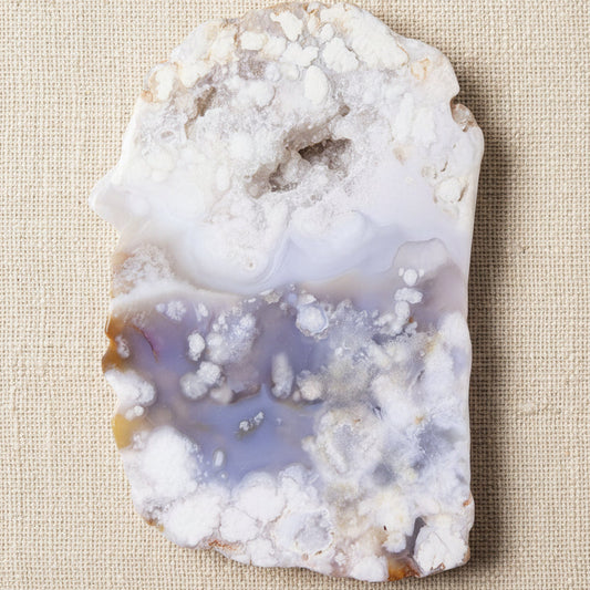A raw flower agate geode slab with a white and lavender-purple banded pattern and crystalline formations