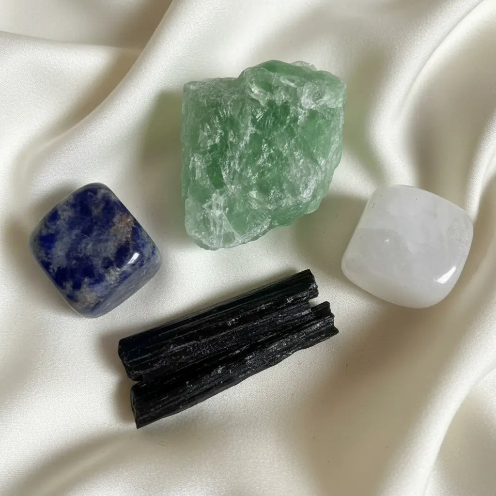 Close up four crystals, Summer Sodalite Carved Cube, Clear Quartz Carved Cube, Black Tourmaline Specimen, Green Fluorite Specimen. Crystals on an off white soft fabric