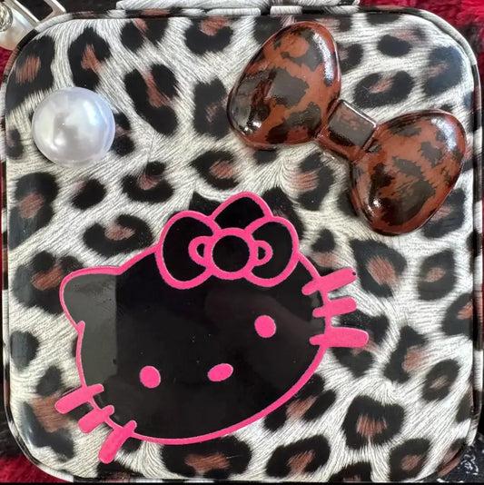 A square compact case featuring a leopard print pattern with a large pink Hello Kitty face graphic, a brown bow embellishment, and a pearl-like bead on a red background.