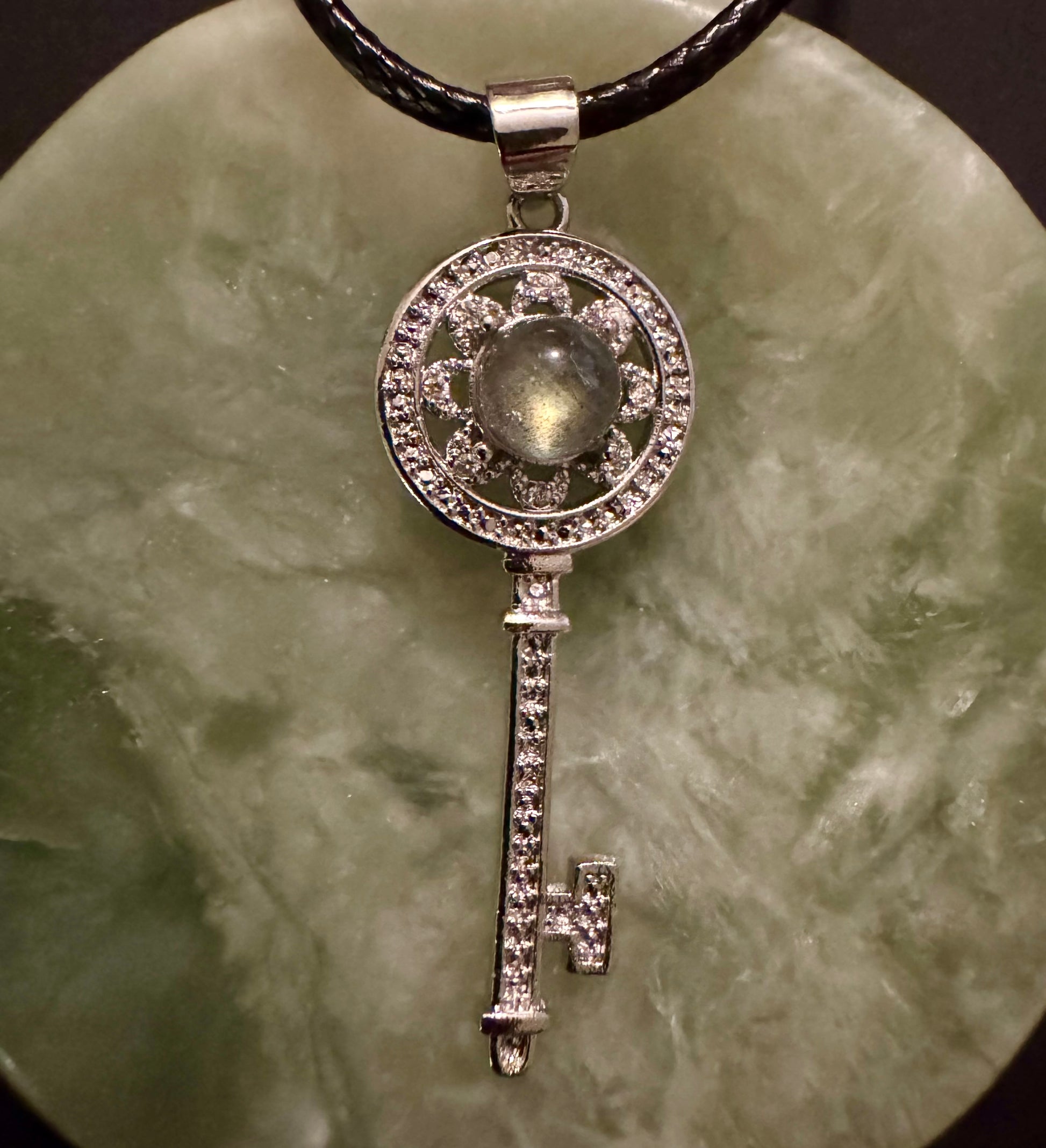 Silver key pendant with a central stone, on a black cord and a mottled green surface.