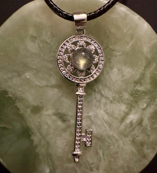 Silver key pendant with a central stone, on a black cord and a mottled green surface.