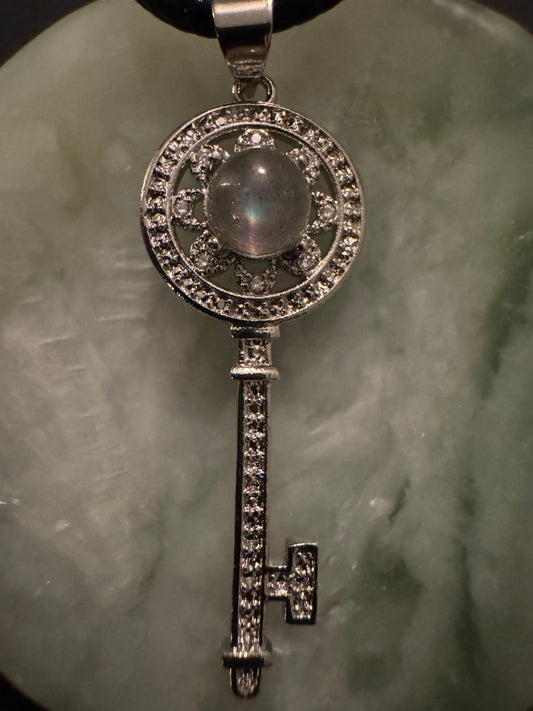 Close-up of a silver, jeweled key pendant necklace on a mottled green surface.