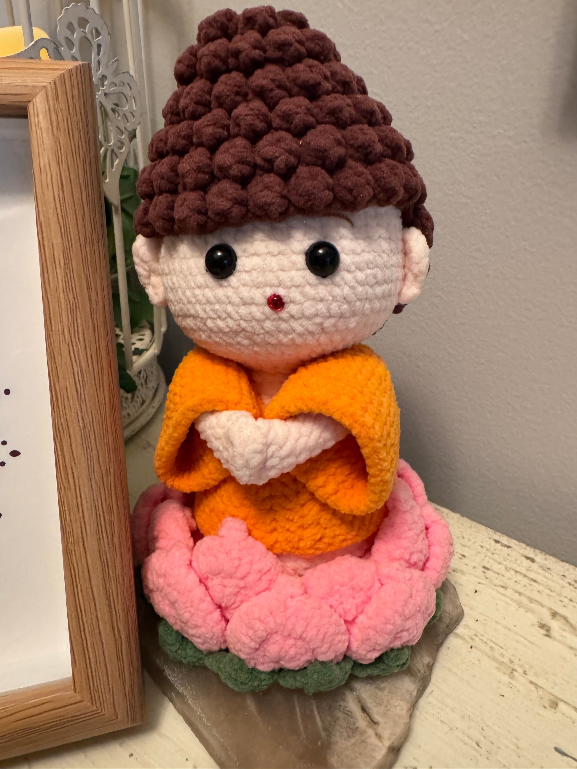 Crocheted doll wearing an orange robe sitting on a pink and green flower.