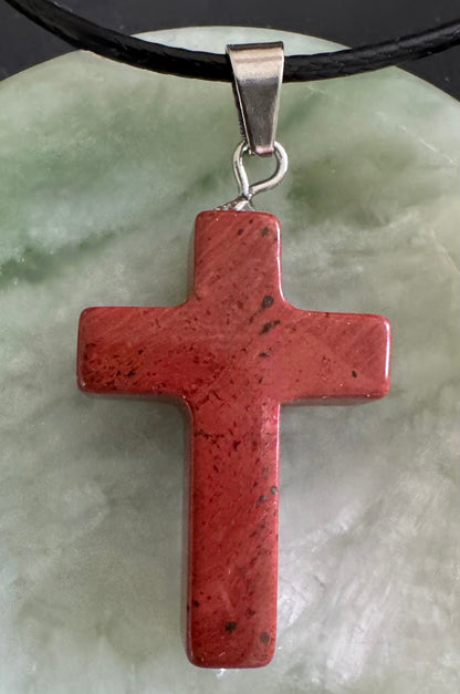A red stone cross pendant with silver clasp and black leather cord on a green stone surface.