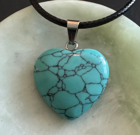 A turquoise heart-shaped stone pendant with black veining hangs from a black leather cord on a light green surface.