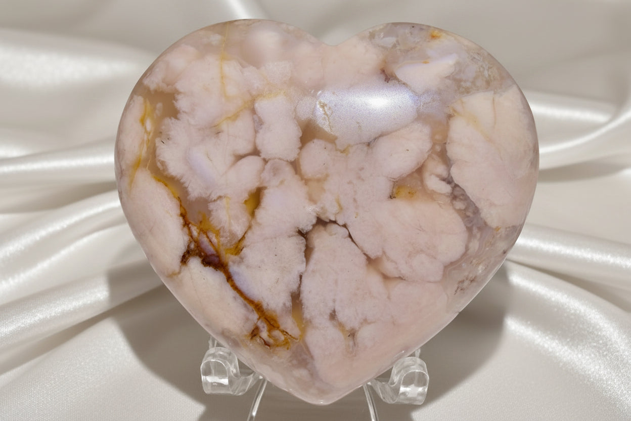 close up of a flower agate hand carved heart crystal is on a small stand with a soft white fabric in the background
