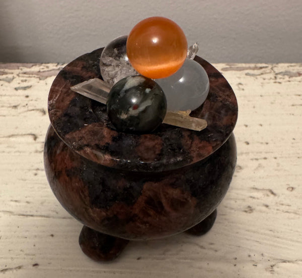 A small decorative pot with a lid and three short legs, topped with several mineral spheres and crystal wands.