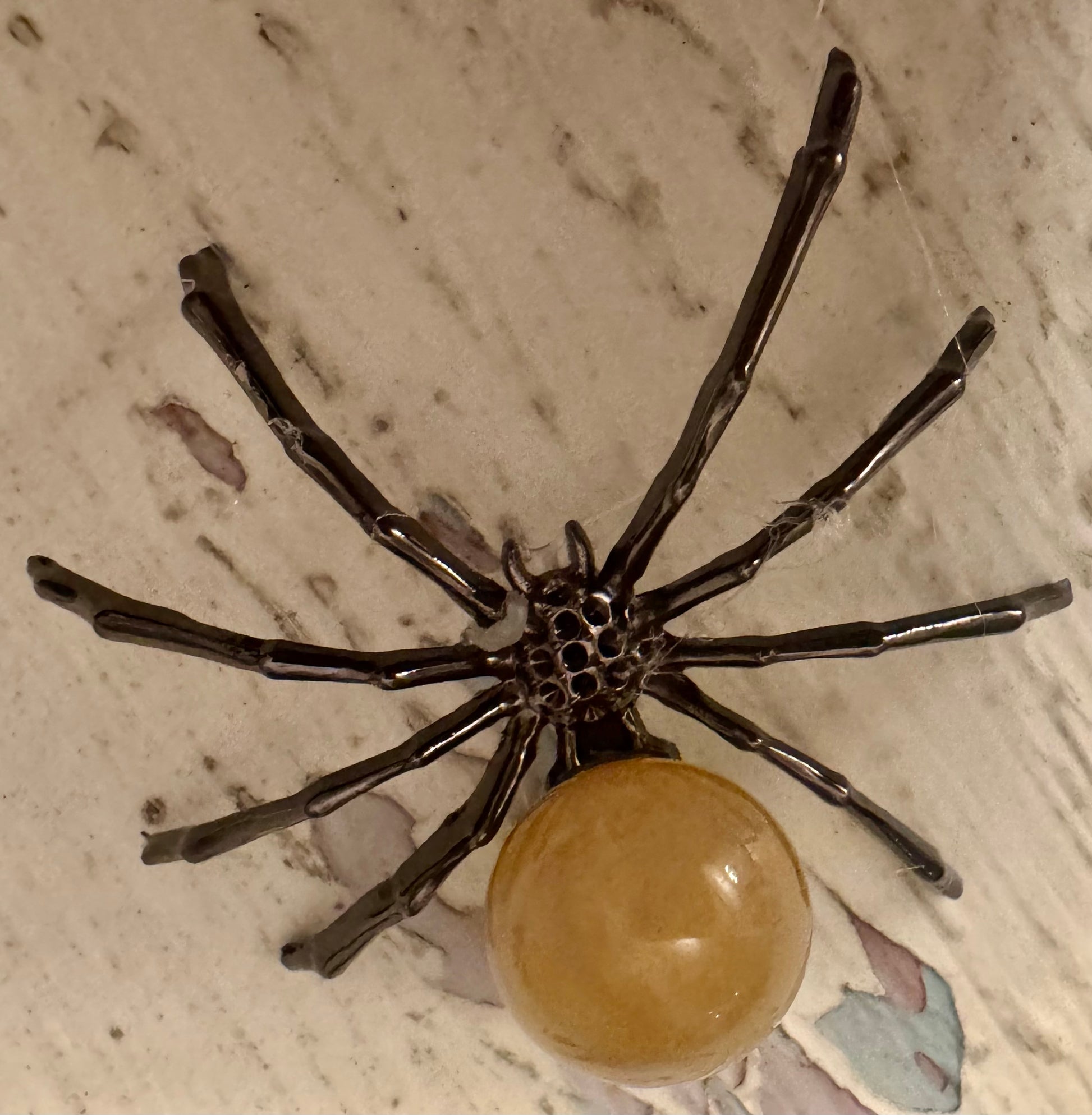 Brooch in the shape of a black spider with an amber-colored round body on a marble surface.