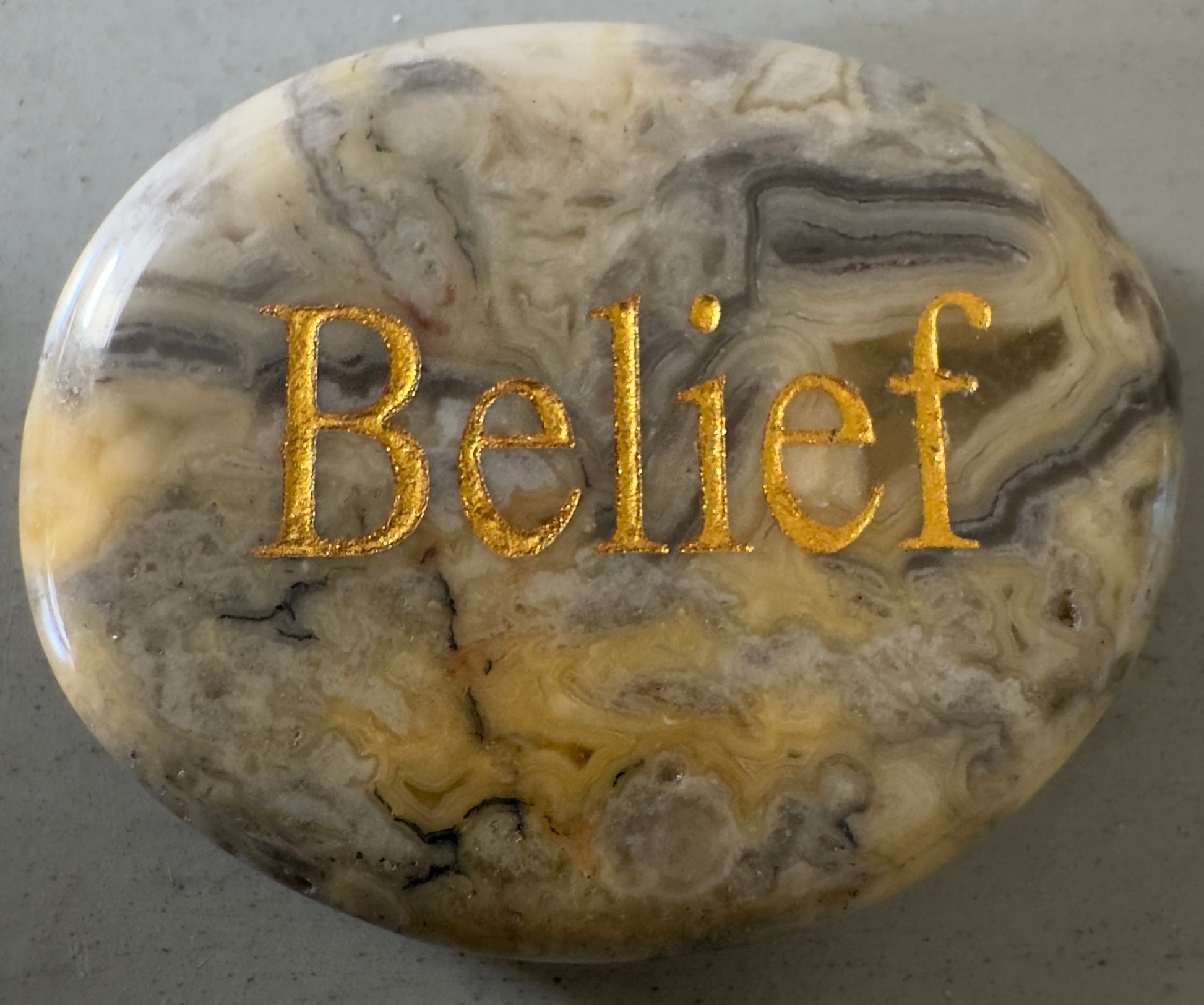Oval stone with layered, swirling gray and yellow patterns; the word "Belief" in gold lettering is centered on the stone. 
