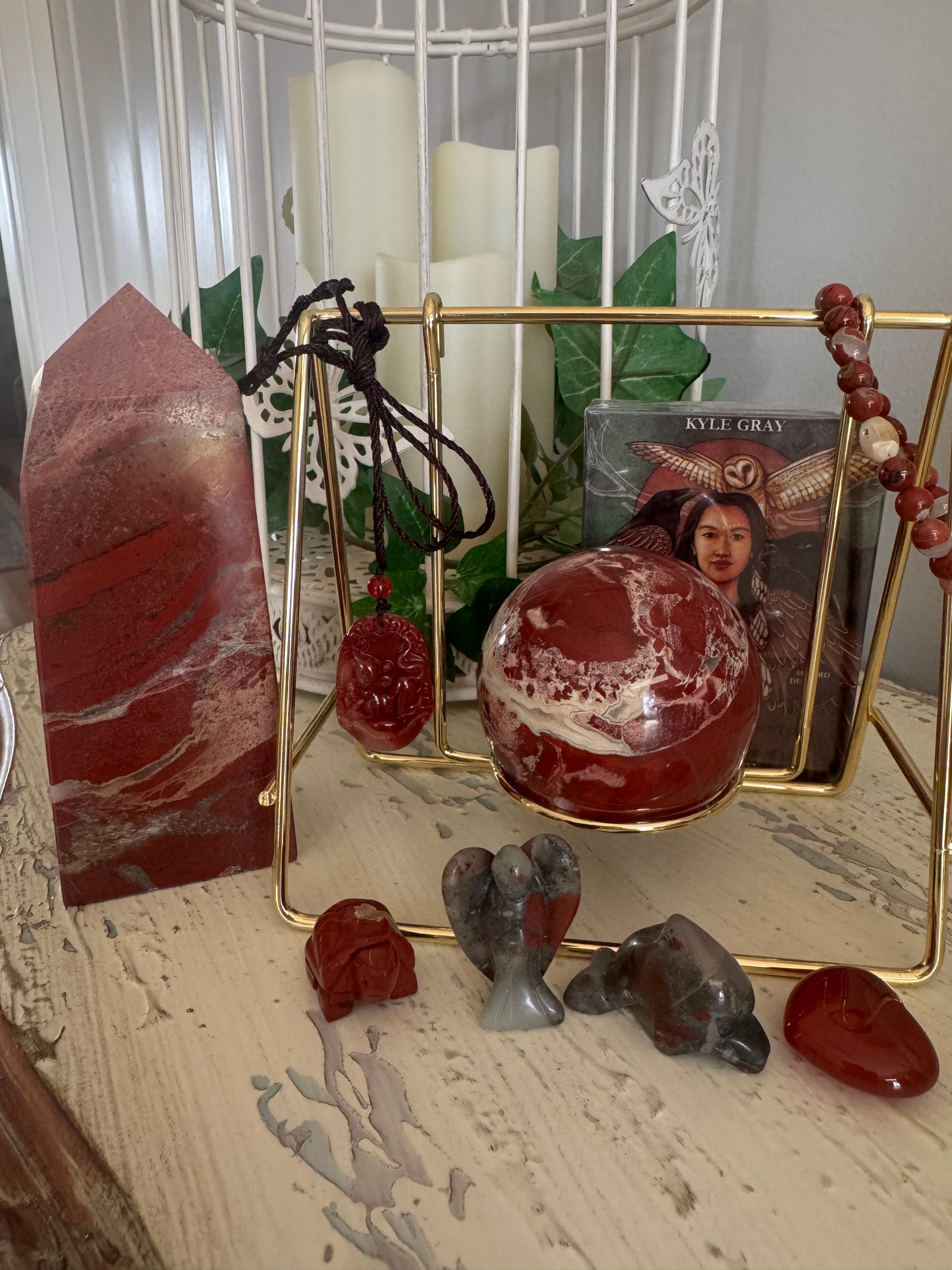 Still life of various red and grey crystals, including a large tower, a sphere, small animal carvings, a pendant, and a strand of beads, arranged around a gold wire stand, with a Kyle Gray oracle card deck in the background, set against a backdrop of white candles and a white decorative birdcage.