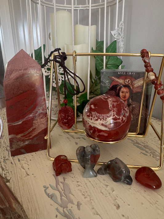 Still life of various red and grey crystals, including a large tower, a sphere, small animal carvings, a pendant, and a strand of beads, arranged around a gold wire stand, with a Kyle Gray oracle card deck in the background, set against a backdrop of white candles and a white decorative birdcage.