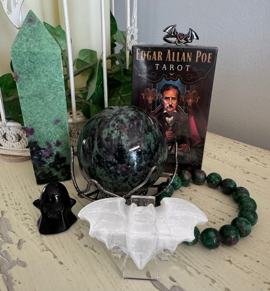 Still life arrangement with an Edgar Allan Poe tarot deck, a crystal ball, a ghost figure, a bat figurine, a green crystal tower, and a beaded bracelet.