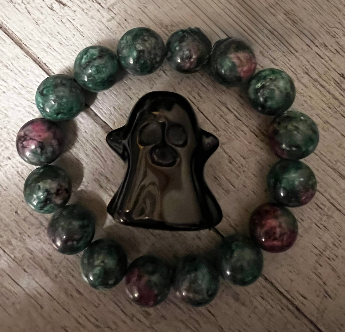 Overhead shot of a glossy black ceramic ghost surrounded by a circle of green and red jasper beads on a gray wood-grain surface.