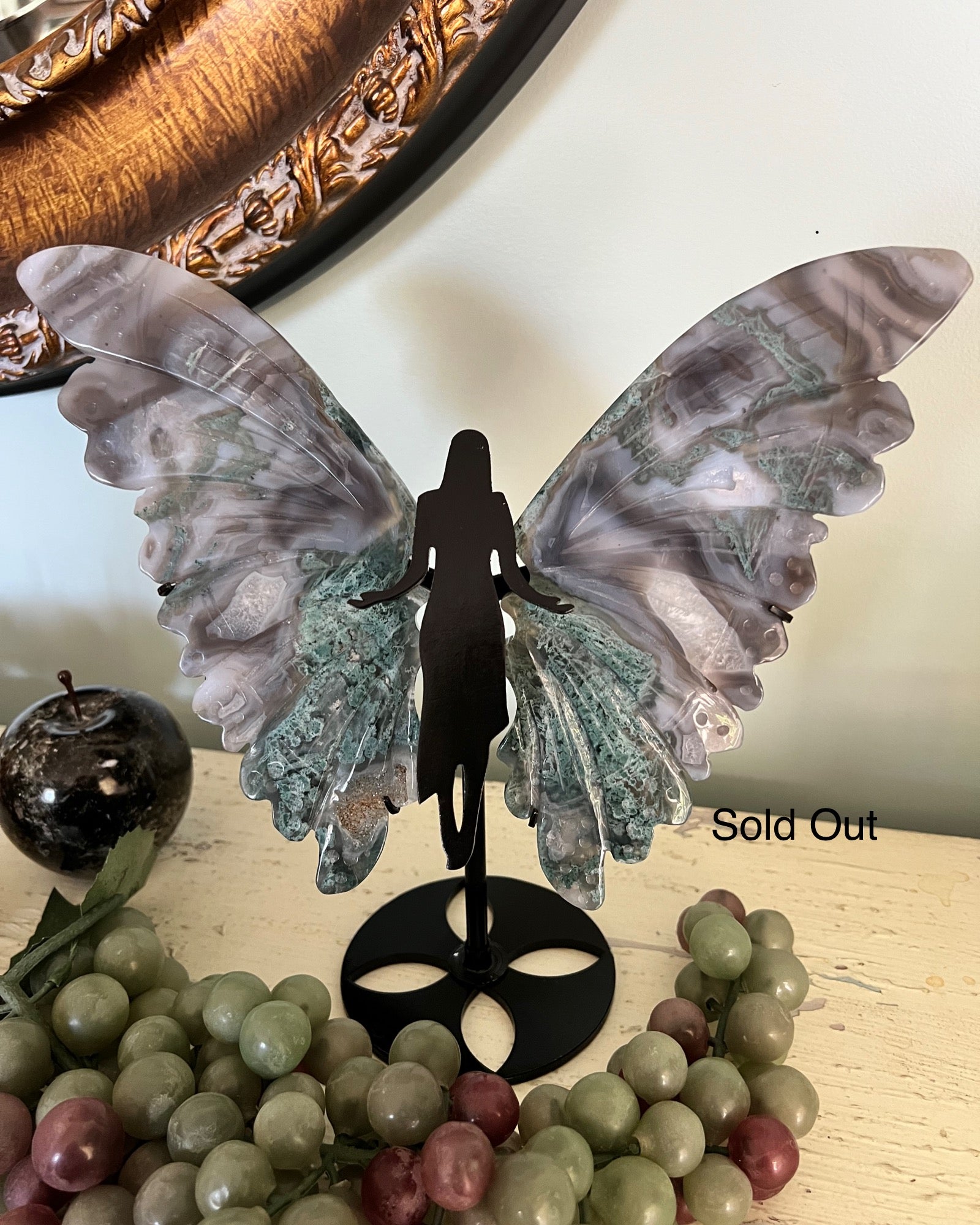 Stone fairy figurine with green and grey wings on a black stand, surrounded by stone grapes and a stone apple.