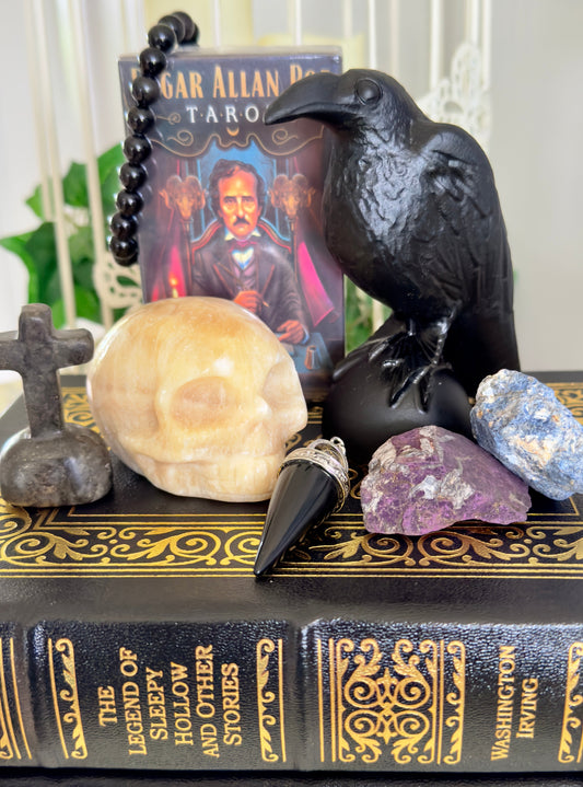 Still life arrangement featuring a book titled "The Legend of Sleepy Hollow," a black raven statue, an Edgar Allan Poe tarot deck, a black bead necklace, a carved skull, a black cone-shaped pendant, a small stone cross, and two chunks of raw crystal. The items are presented against a blurry background.
