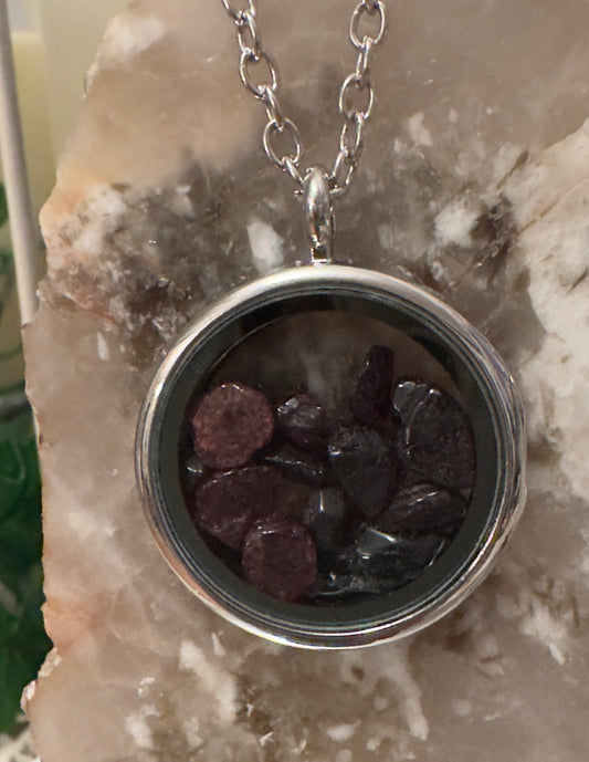 A silver necklace with a pendant containing dark red and black stones, displayed against a marbled stone surface.
