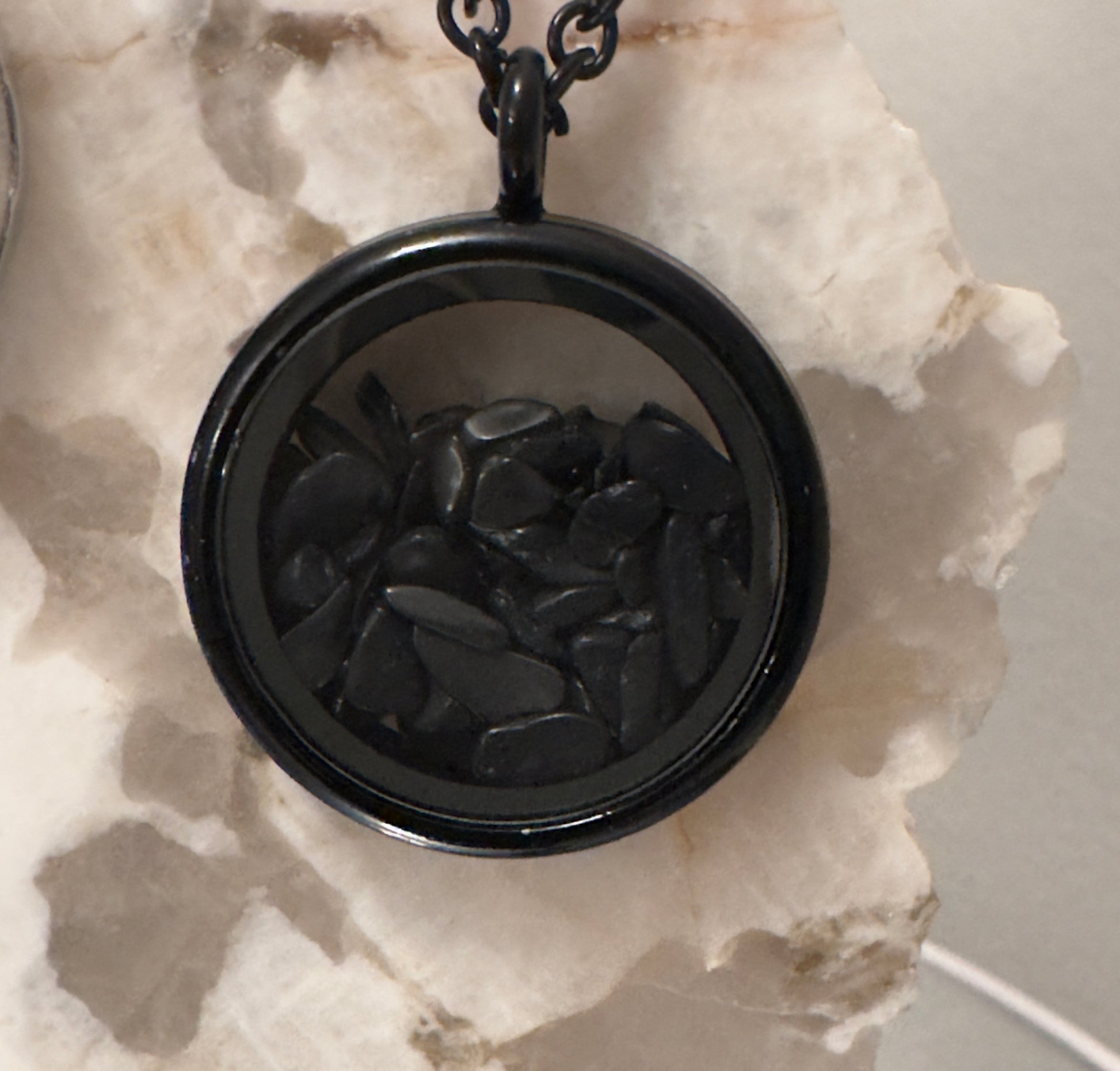 Black circular locket pendant with small black stones inside, hanging on a chain, resting on a light-colored stone surface.
