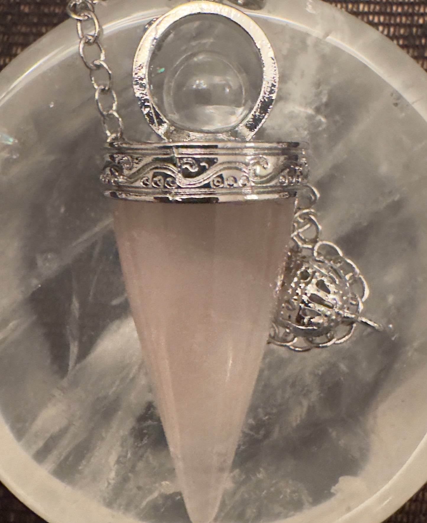 A rose quartz pendulum with a silver chain and a decorative silver cage rests on a cloudy gray and white surface.