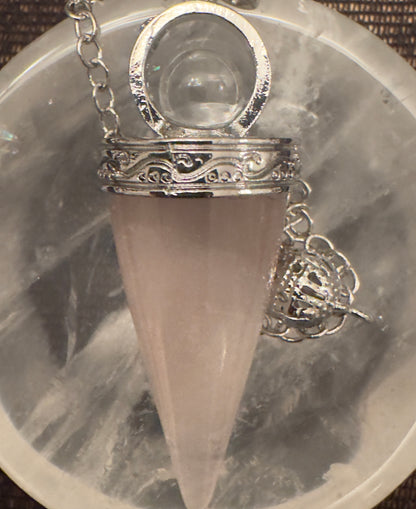 A rose quartz pendulum with a silver chain and a decorative silver cage rests on a cloudy gray and white surface.