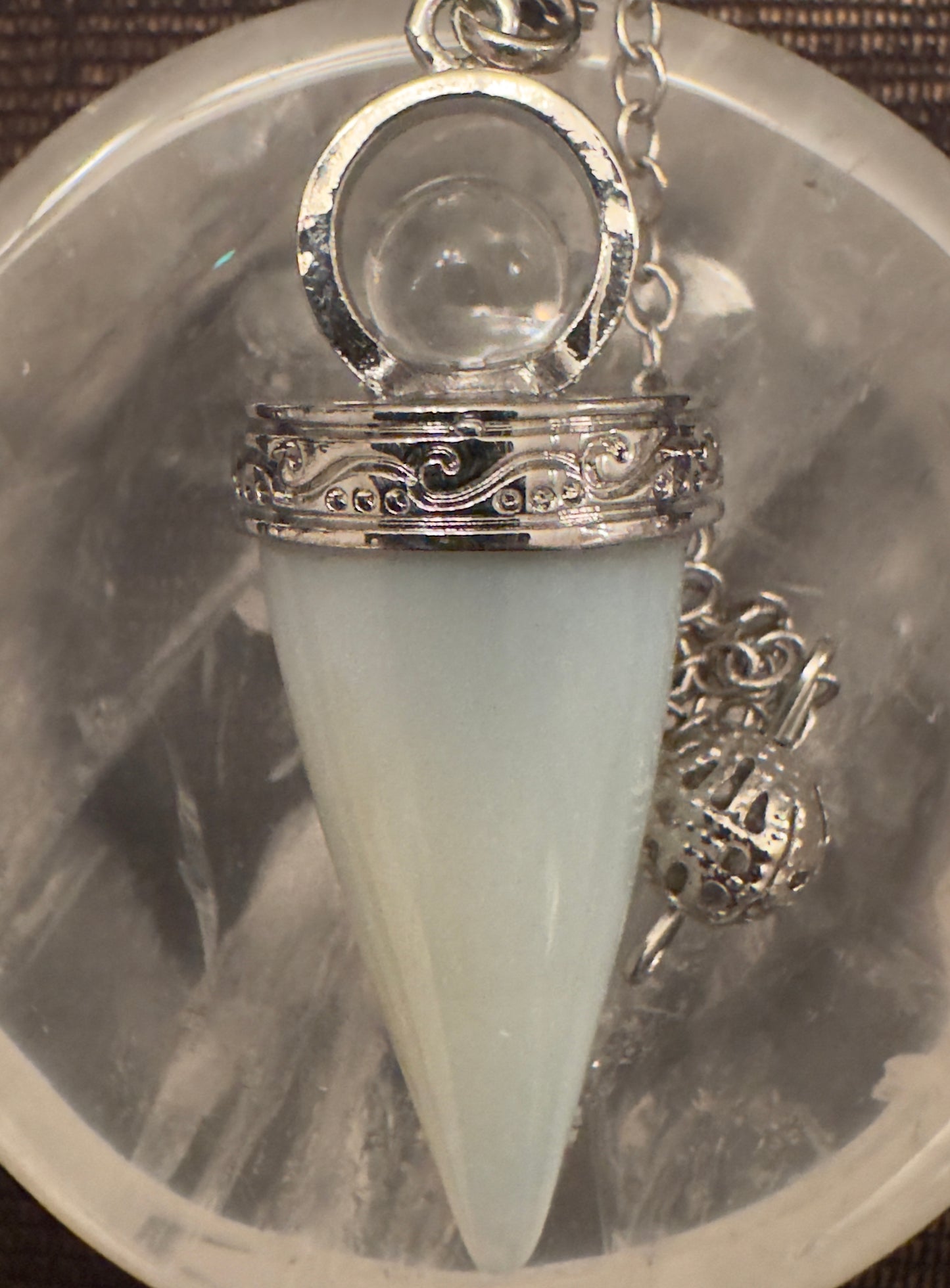 Close-up of a silver pendulum with a light green pointed crystal hanging over a geode-like surface.