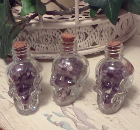 Three clear skull-shaped bottles filled with purple crystals, sealed with corks.
