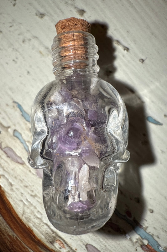 A clear skull-shaped bottle with a cork stopper, filled with amethyst stones, on a patterned surface.