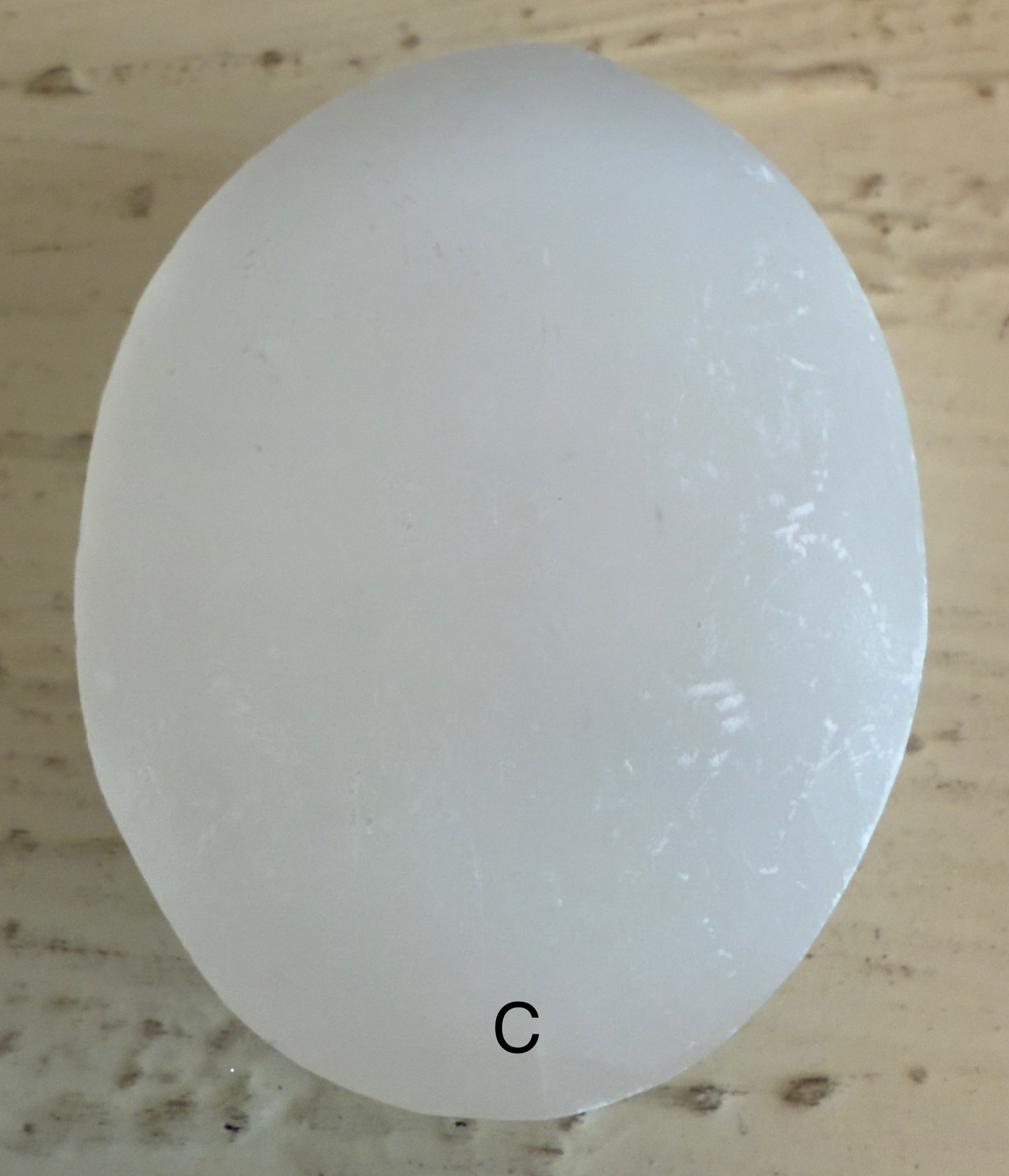 Oval, translucent white stone with the letter "C" at the bottom on a textured tan surface.
