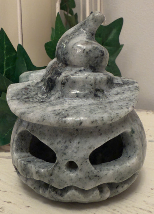 A stone pumpkin with a witch's hat sitting on a table with green leafy plants in the background.