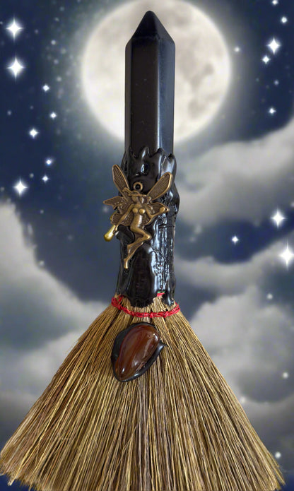 A witch's broom with a black crystal on top, adorned with a brass fairy and a brown stone against a night sky backdrop.