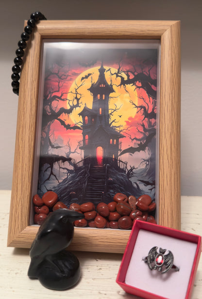 Framed print of haunted house, red stone fill, black beaded necklace, raven figurine, and vampire bat ring.