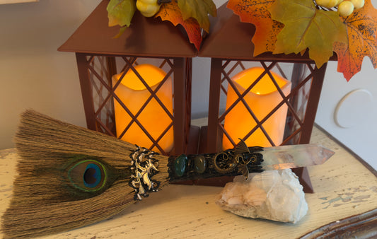 Two brown lanterns with autumn leaves, a broom with a peacock feather, a witch charm, and a decorative dragon-encrusted wand over a quartz cluster.