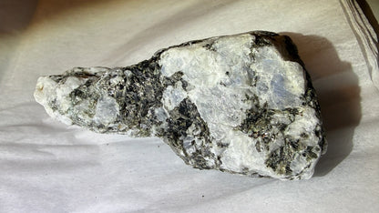 A raw mineral specimen with a mix of white and dark gray crystalline texture, resting on a white fabric background.