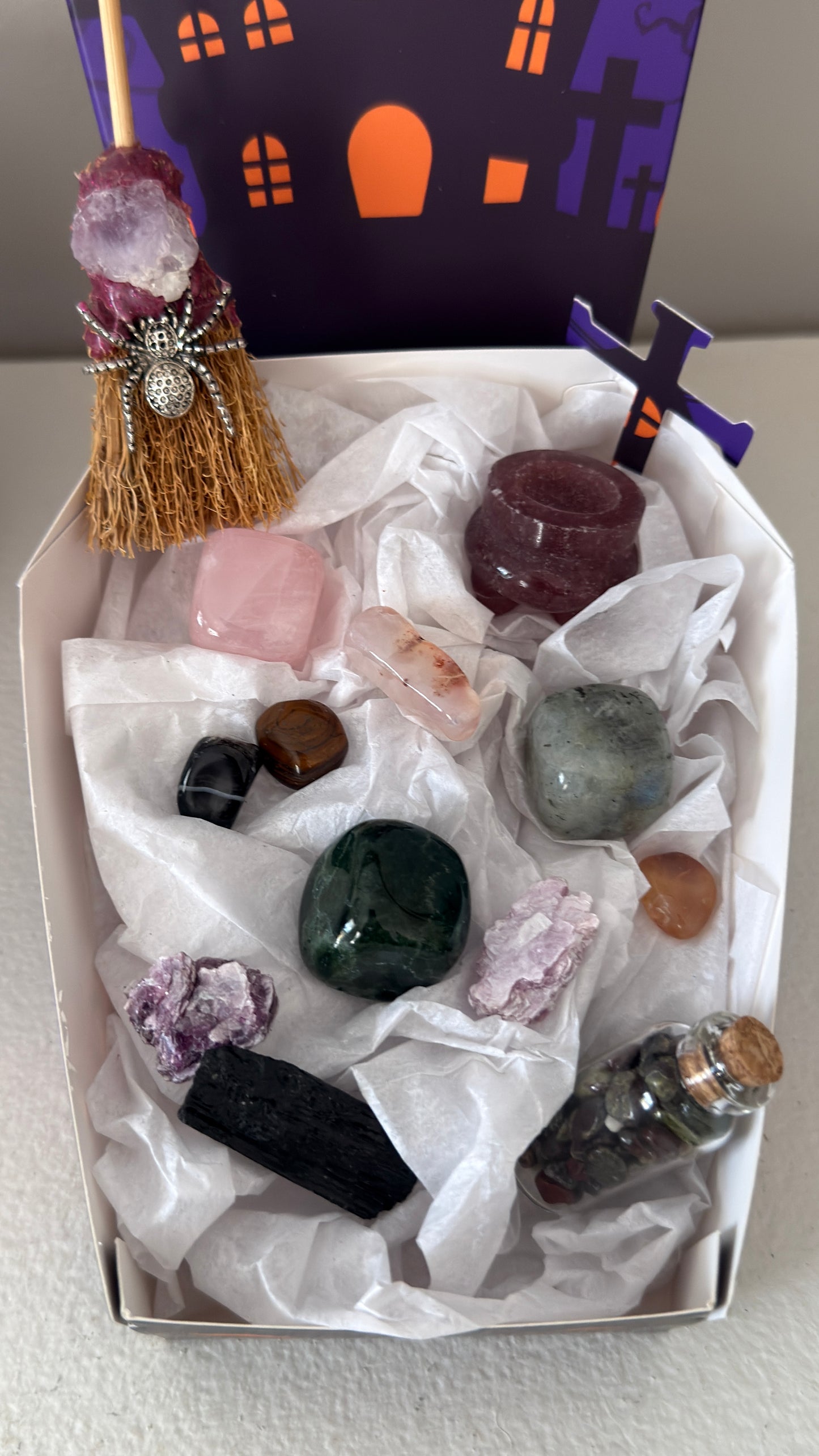 close up photo of all included pieces in bundle. rose quartz, green jasper and labradorite cubes. Dragon’s Blood wish bottle, Black Tourmaline Raw, Strawberry Quartz carved cauldron, assorted tumbles, witches broom with amethyst and silvery spider charms in a paper vessel purple and orange shaped like a coffin decorated in halloween design