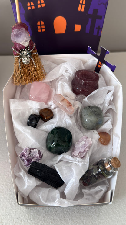 close up photo of all included pieces in bundle. rose quartz, green jasper and labradorite cubes. Dragon’s Blood wish bottle, Black Tourmaline Raw, Strawberry Quartz carved cauldron, assorted tumbles, witches broom with amethyst and silvery spider charms in a paper vessel purple and orange shaped like a coffin decorated in halloween design