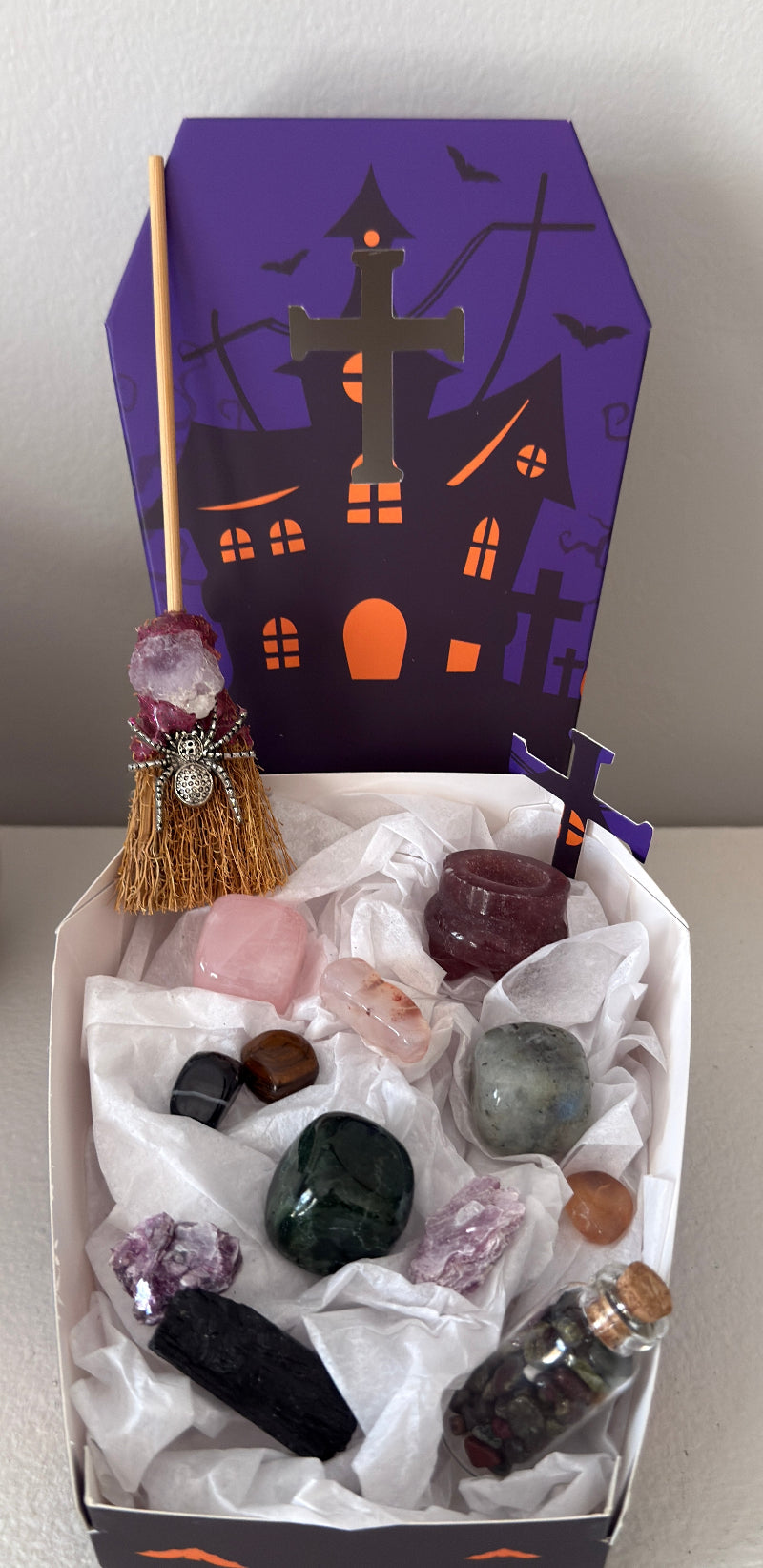 photo of actual pieces and complete set bundle Witch’s broom, assorted crystal tumbles, strawberry quartz cauldron, green jasper, labradorite and green jasper cubes, black tourmaline raw stone, wish bottle with dragon’s blood chips, tumbles and pieces of mica. Background shows lid to specialty paper box included. Coffin shaped two piece box, purple and orange decorated with a halloween house, bats and tomb stones